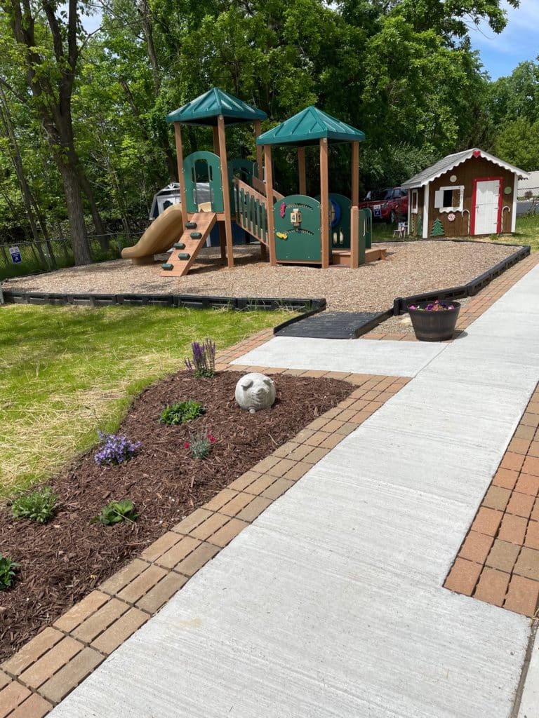 Photograph of the backyard at The Children's Center of Medina County shows brick pavers lining both sides of the sidewalk. There is a landscaped garden with newly planted flowers along the pavers on the left. In the background, there is a playground and a gingerbread-style playhouse.