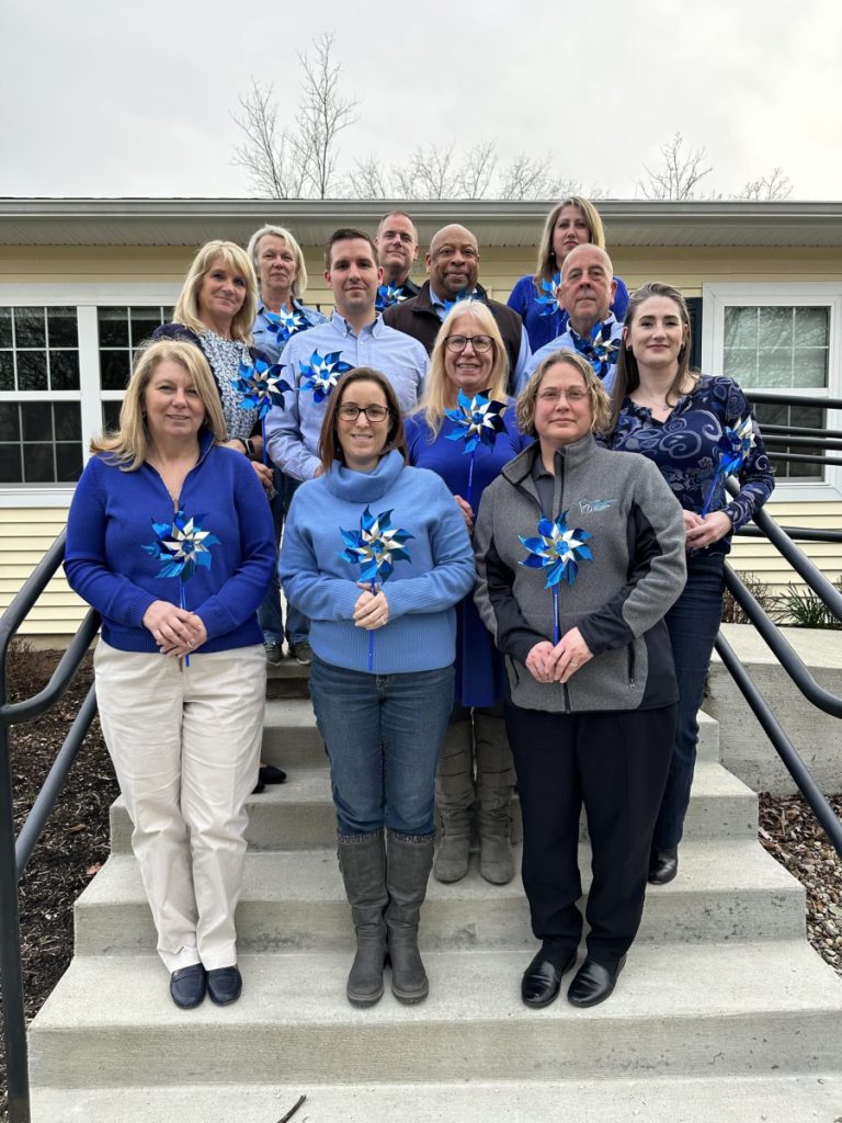 Twelve men and women serving on The Children's Center Board of Directors stand on the steps in front of a yellow building. They are wearing blue clothing in support of Child Abuse Prevention Month