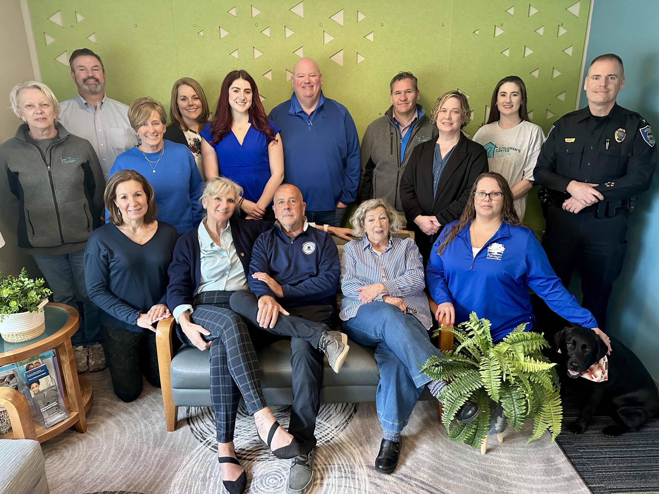 Twelve men and women serving on The Children's Center Board of Directors stand on the steps in front of a yellow building. They are wearing blue clothing in support of Child Abuse Prevention Month