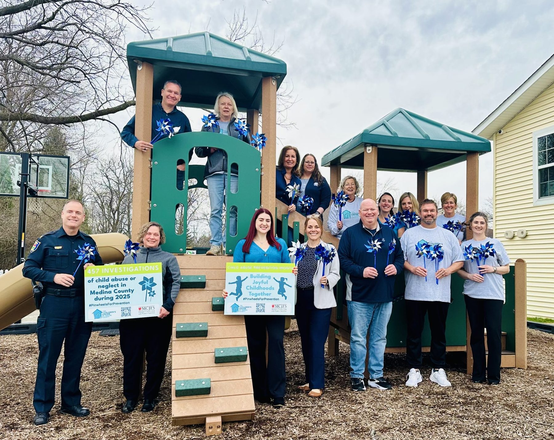 Twelve men and women serving on The Children's Center Board of Directors stand on the steps in front of a yellow building. They are wearing blue clothing in support of Child Abuse Prevention Month