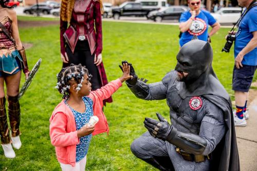 A man dressed in a Batman costume squats down to give a young girl with beads in her hair a high-five