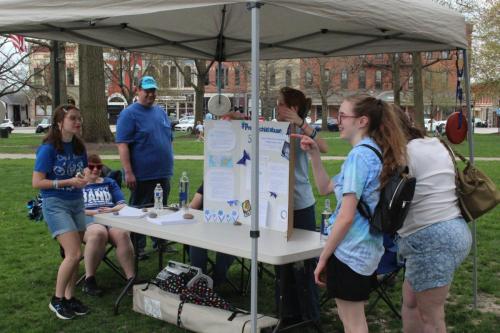 A group of teens and adult girl scouts stand at a table behind a display board