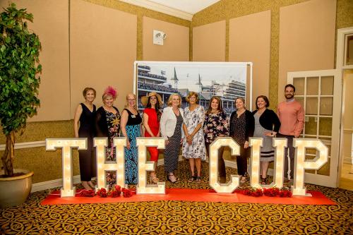 A group of adults standing in front of a Kentucky Derby themed backdrop with light up letters in front of them spelling out THE CUP