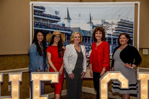 A group of five women standing in front of the Kentucky Derby themed backdrop with a portion of light up letters in front of them