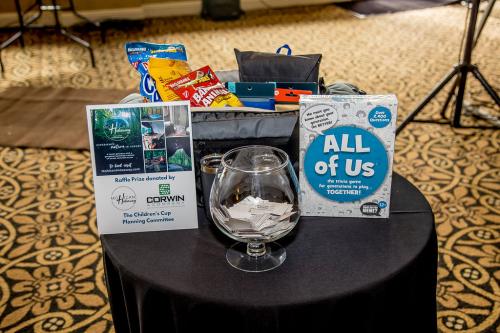 A small round table with black table cloth has two signs with information about the Mohican Hideaway Raffle. There is a round glass bowl partially filled with raffle tickets in the front. Behind these items is a black duffle bag with various snacks and items for the raffle winner