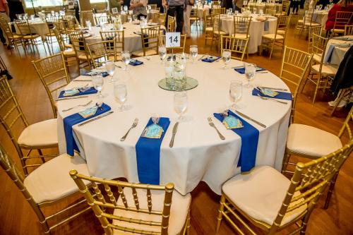 A round table with white tablecloth, blue napkins, gold chairs, and a clear centerpiece with floating candles on a round mirror