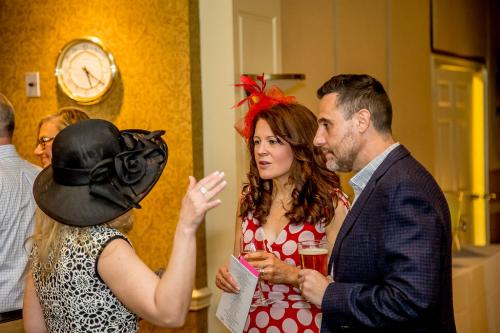 A woman in a black hat gesturing with her hand while talking to a woman in a red dress with a red feather hat and a man in a dark suit jacket