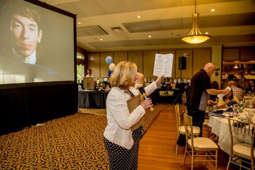 A woman in a white blazer and black and white polka dot pants speaking into a microphone she is holding with one hand while holding up an open event program in the other hand