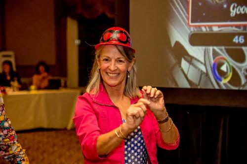 A woman in a pink blazer and polka dot shirt wearing a red plastic jockey hat pretending to giddy-up